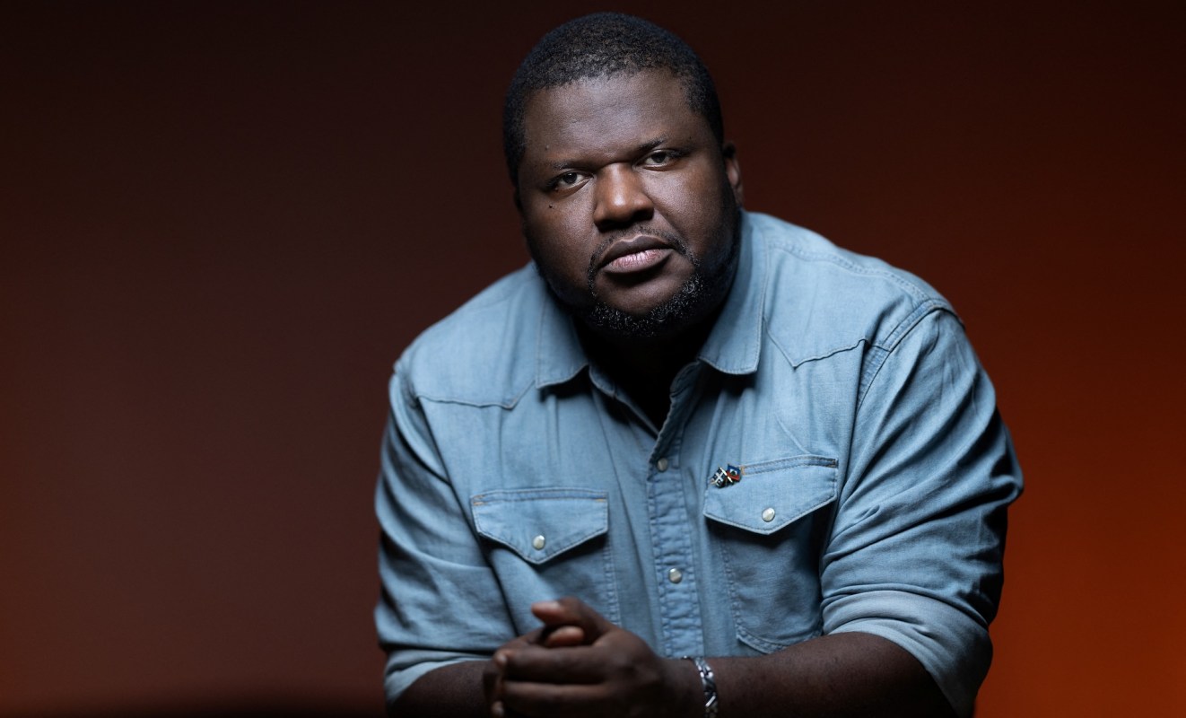Writer Thelyson Orelien, wearing a denim shirt, sits with hands clasped in front of a dark orange backdrop.
