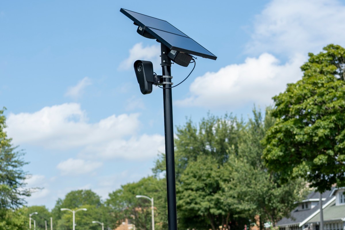 A Flock Safety camera with a solar panel mounted on a pole with a camera and speaker in an outdoor setting.
