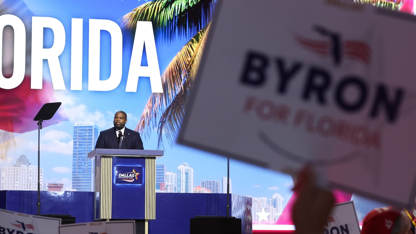 U.S. Rep. Byron Donalds addresses an audience with a large backdrop displaying "FLORIDA" and signs for a political campaign.