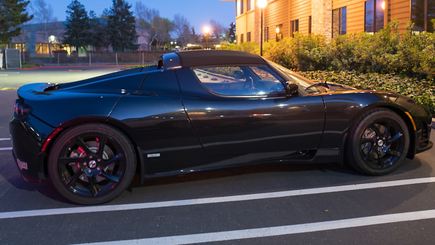 A black Tesla Roadster parked in a lot near a building at dusk.