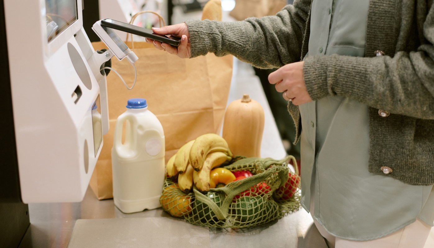 A person scans a smartphone at a self-checkout with groceries nearby, including milk and produce.