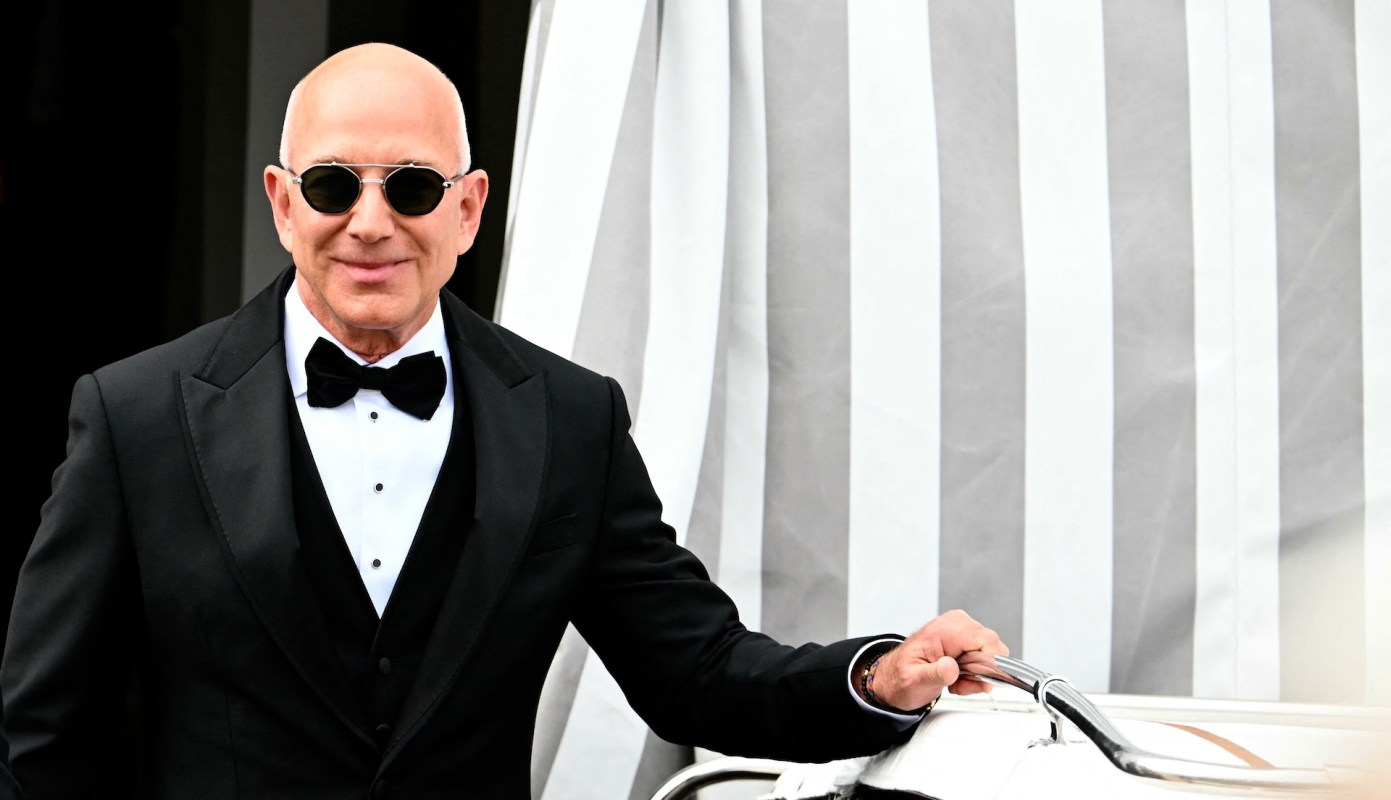 Jeff Bezos in a tuxedo and sunglasses stands next to a boat with a striped backdrop.