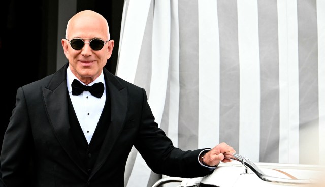 Jeff Bezos in a tuxedo and sunglasses stands next to a boat with a striped backdrop.