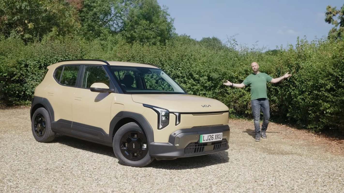 A man stands beside a beige Kia SUV in a natural setting with greenery in the background.