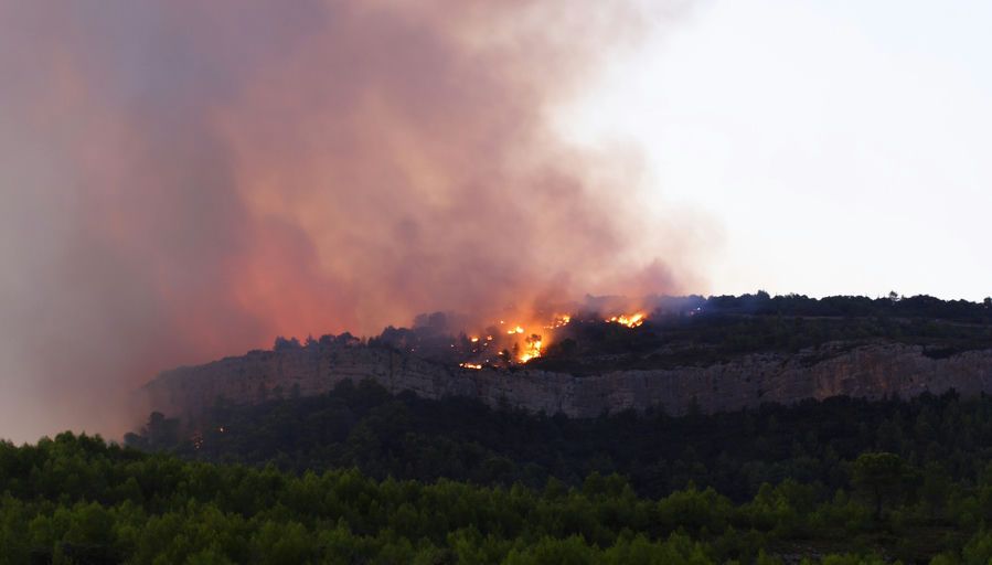A forest fire burns on a hillside, with thick smoke rising into the sky.