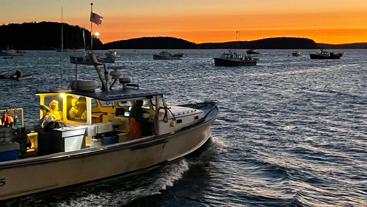 A fishing boat navigates through water at sunset, with other boats visible in the background.