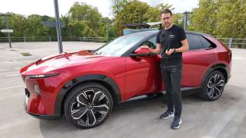 A man stands beside a red SUV on a parking lot with trees in the background.