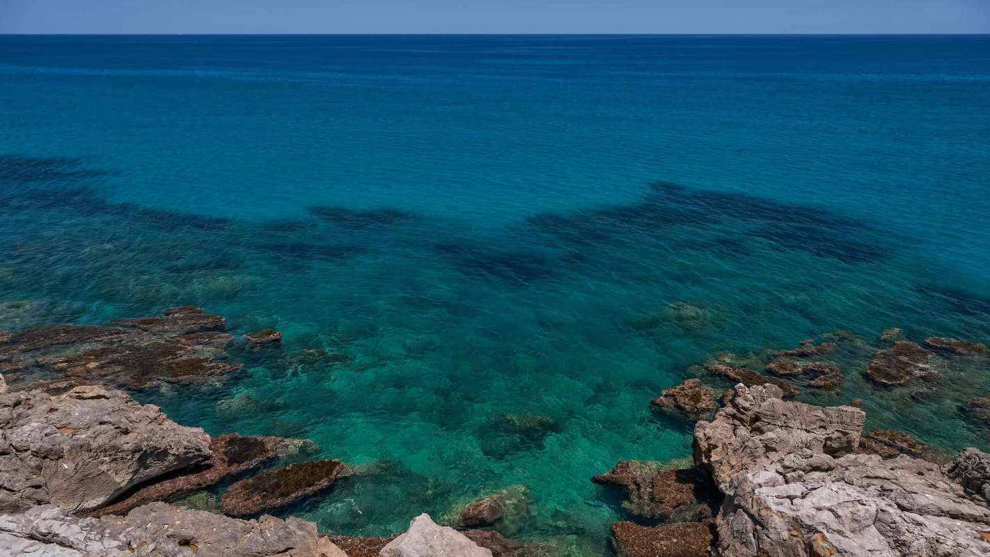 Clear turquoise water with visible rocks below the surface and a calm sea in the background.