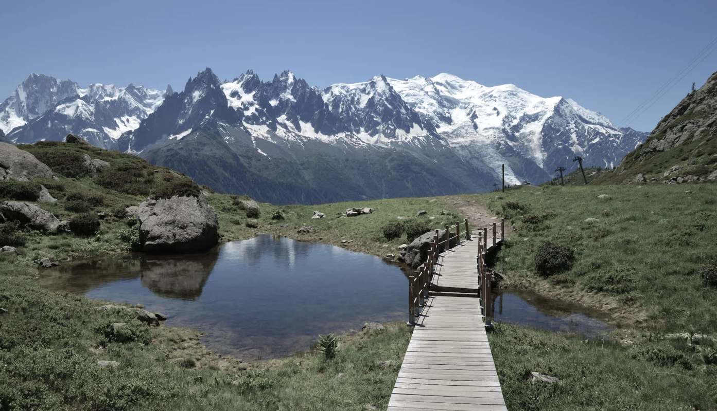 A wooden pathway leads across a grassy area with a pond, set against a backdrop of snow-capped Mont Blanc.