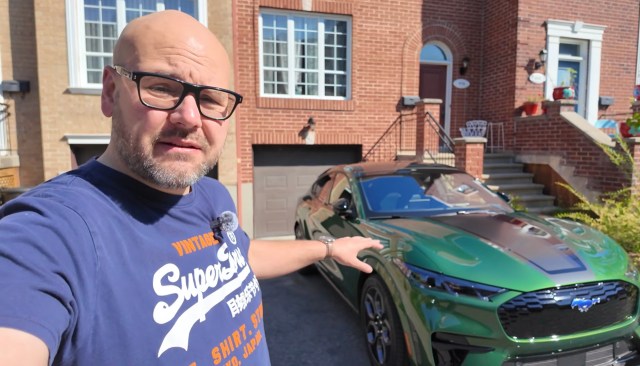 A man stands next to a green car in a residential driveway.