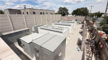 A large outdoor area with electrical equipment, including battery storage containers and machinery near a fenced wall.