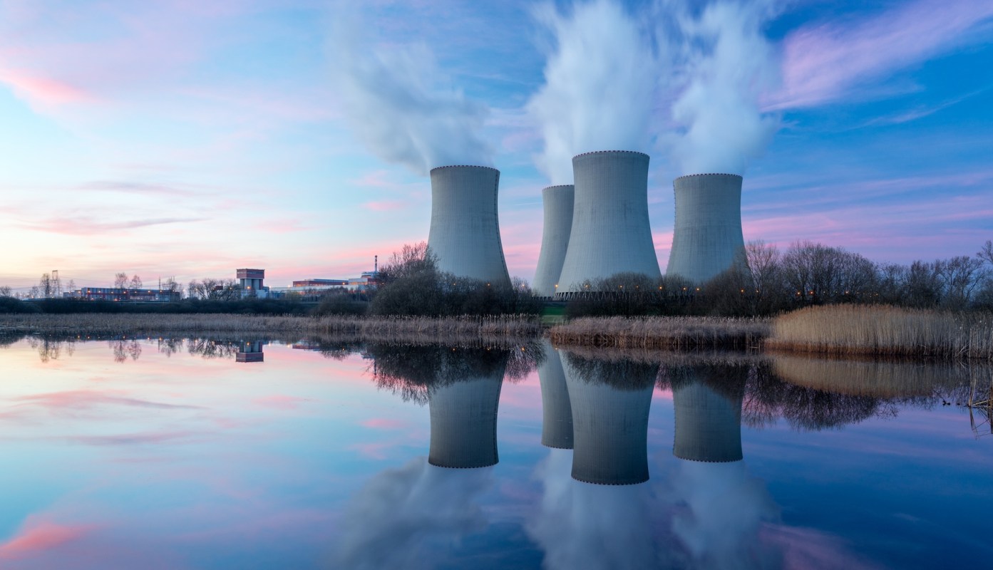 Cooling towers and a power plant reflected in a calm body of water during sunset.