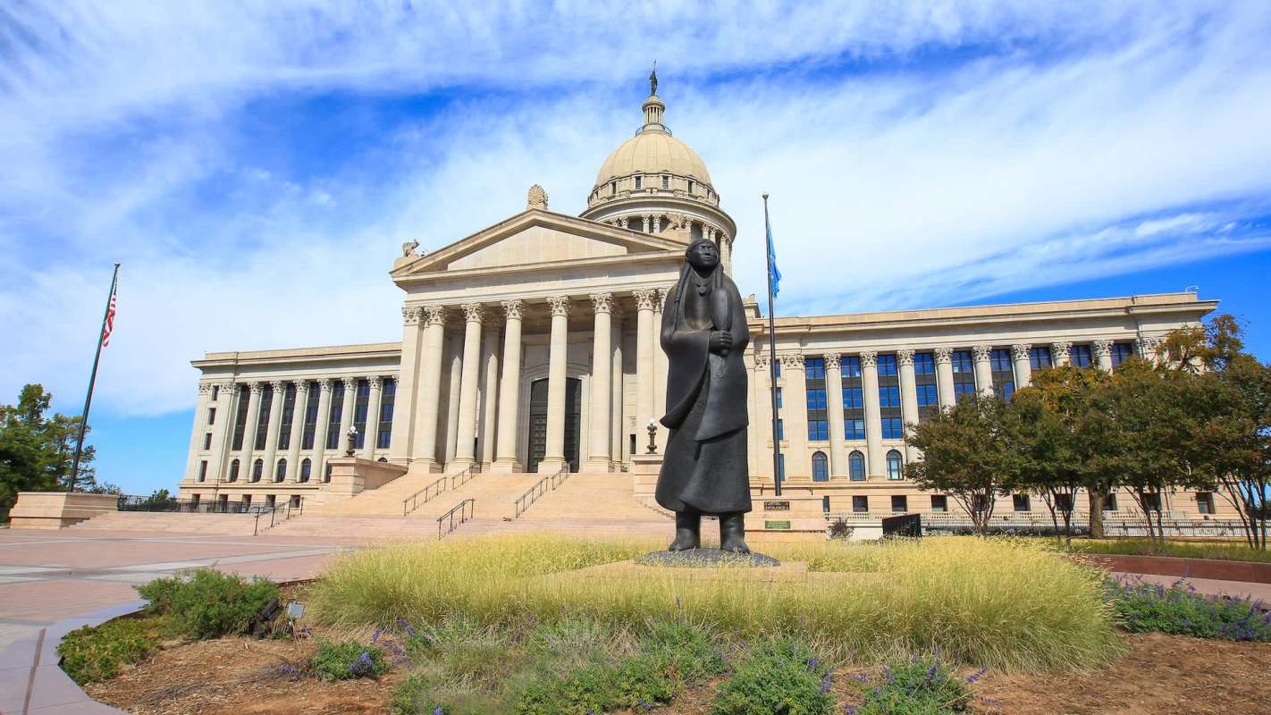 A statue stands in front of the Oklahoma State Capitol with a dome and columns, surrounded by grass and trees.