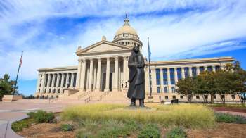 A statue stands in front of the Oklahoma State Capitol with a dome and columns, surrounded by grass and trees.