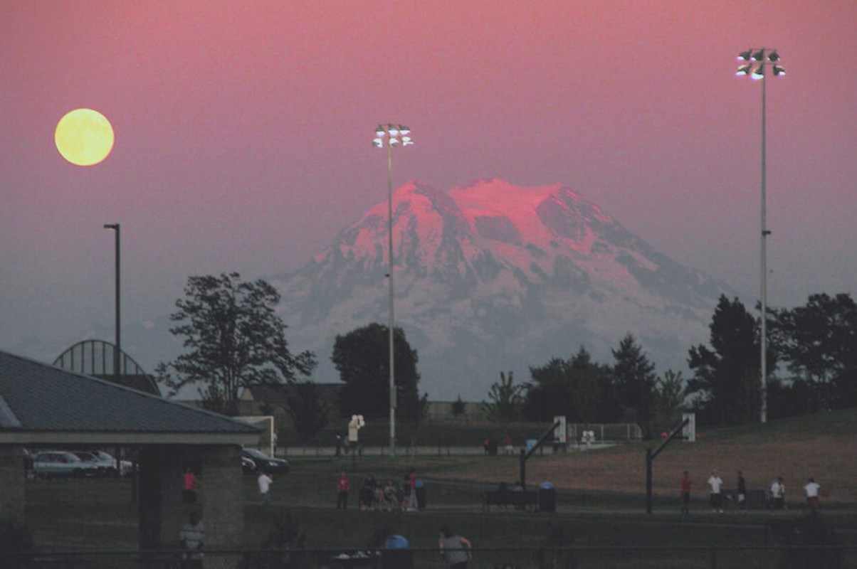 A full moon rises over a mountain with sports fields and trees in the foreground.