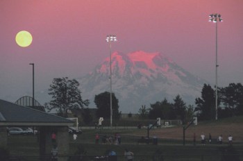 A full moon rises over a mountain with sports fields and trees in the foreground.