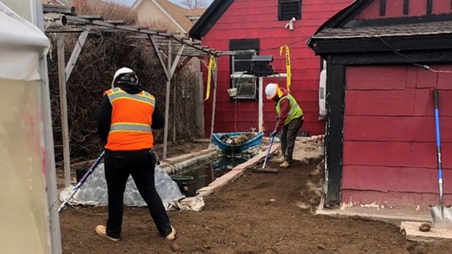 Two workers in safety vests and helmets are cleaning up a yard near a red building with a small boat and a water feature.