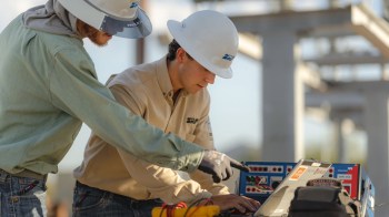 Two workers in hard hats examine a laptop and electrical equipment at a construction site.