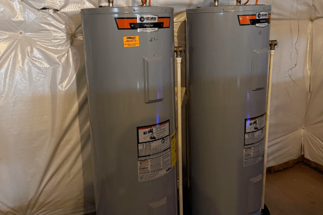 Two gray water heaters are installed side by side in a basement setting.
