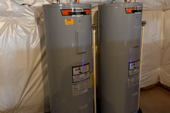 Two gray water heaters are installed side by side in a basement setting.
