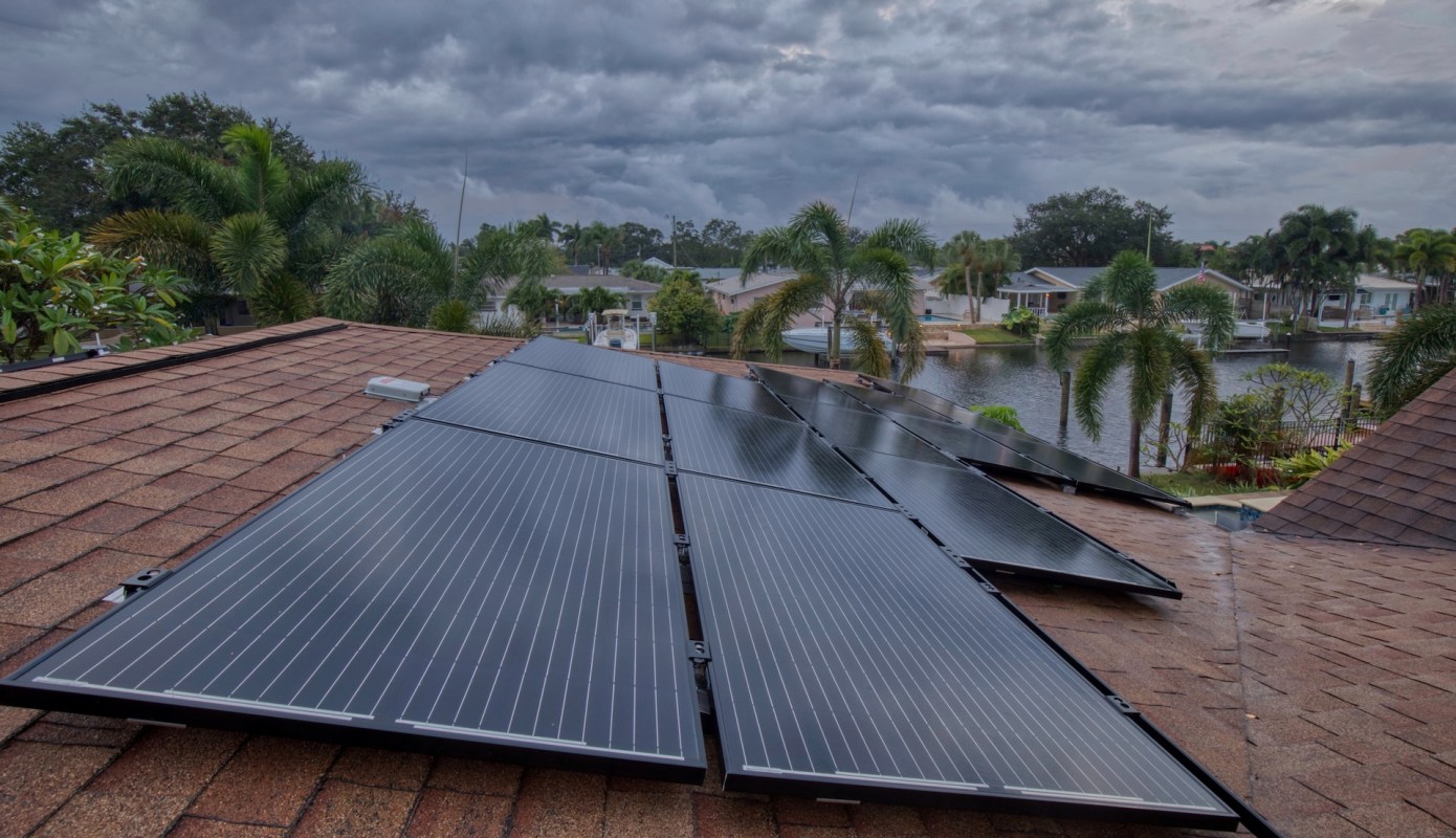 Solar panels installed on a roof with houses and palm trees visible in the background.