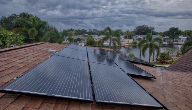 Solar panels installed on a roof with houses and palm trees visible in the background.