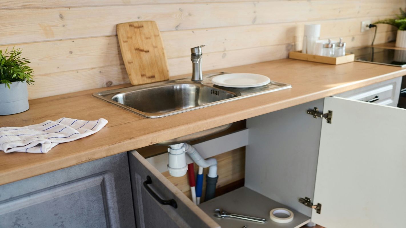A kitchen countertop with a sink, cutting board, plates, and an open cabinet showcasing plumbing and various tools.