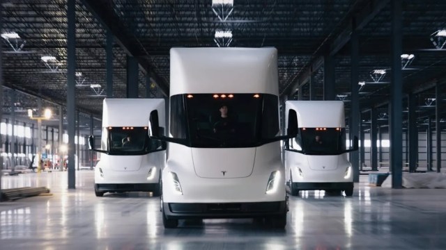 Three Tesla Semitrucks positioned indoors with a driver visible in the front vehicle.