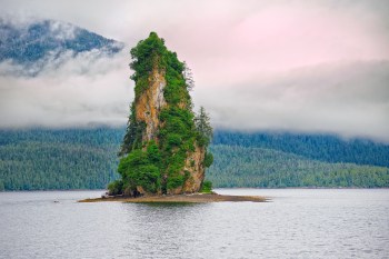 A rock formation covered in greenery rises from a body of water, surrounded by a forest and foggy mountains.
