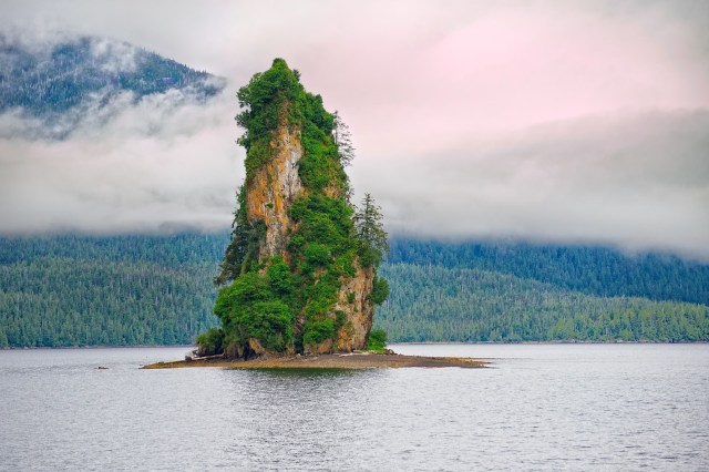 A rock formation covered in greenery rises from a body of water, surrounded by a forest and foggy mountains.