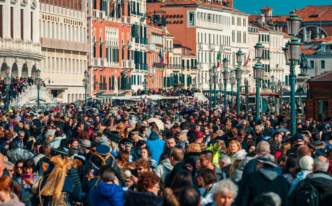 A large crowd of people fills a Venice, Italy, street alongside historic buildings and lampposts.