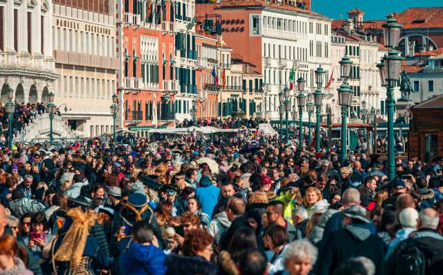 A large crowd of people fills a Venice, Italy, street alongside historic buildings and lampposts.