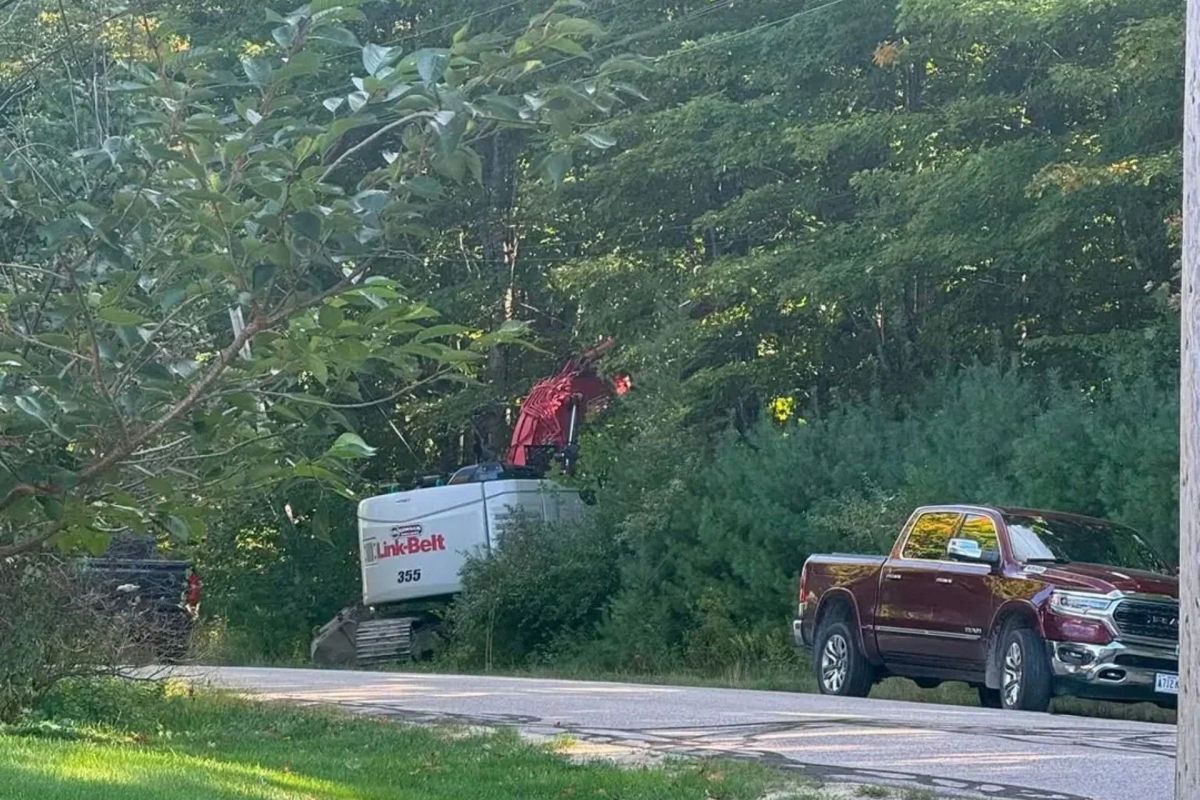 A construction vehicle parked near a road with trees and a red pickup truck in the background.