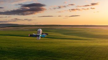 A drone hovers above a grassy landscape at sunset.