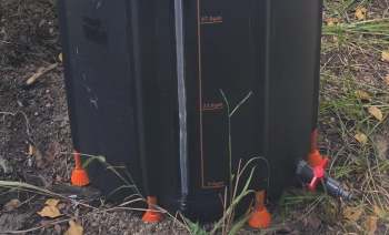 A black rain barrel with an attached hose and measurement markers is placed on the ground among grass and leaves.
