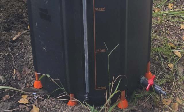 A black rain barrel with an attached hose and measurement markers is placed on the ground among grass and leaves.