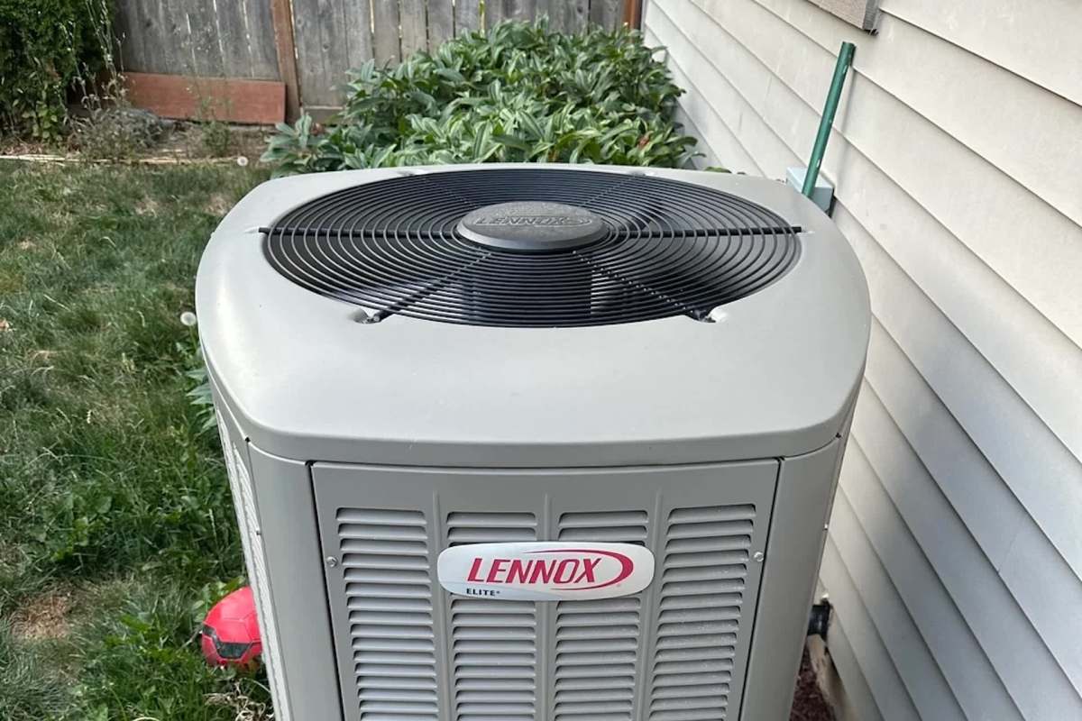 An outdoor air conditioning unit with a black fan on top, surrounded by greenery and a wall.