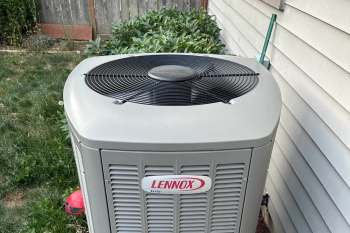 An outdoor air conditioning unit with a black fan on top, surrounded by greenery and a wall.
