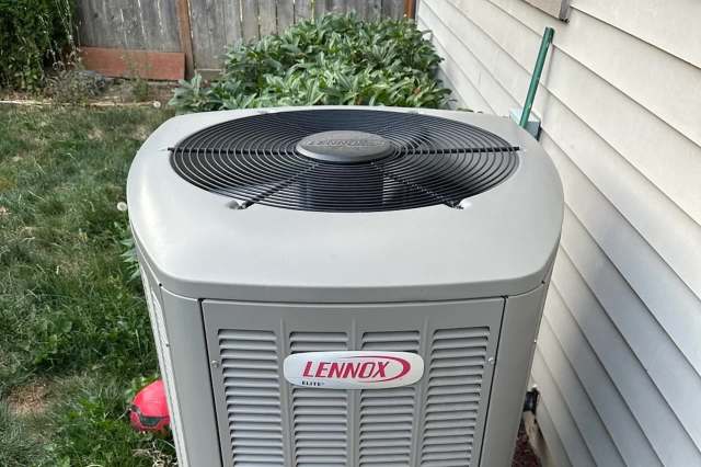 An outdoor air conditioning unit with a black fan on top, surrounded by greenery and a wall.