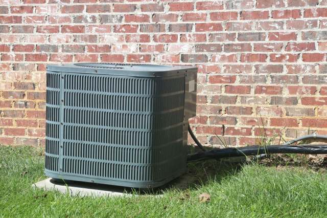 An air conditioning unit beside a brick wall with grass and electrical wiring nearby.