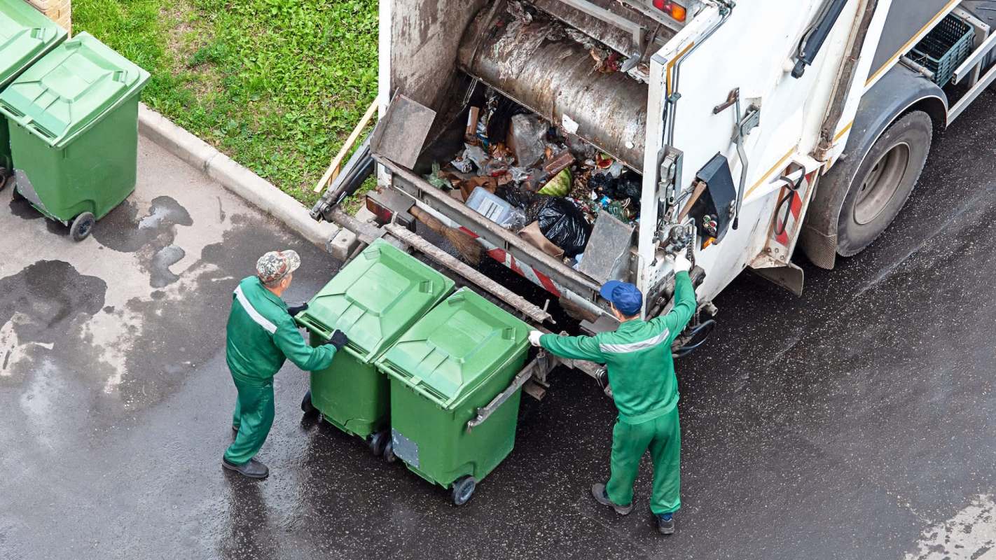 Two workers unload green trash bins into a garbage truck in a paved area.