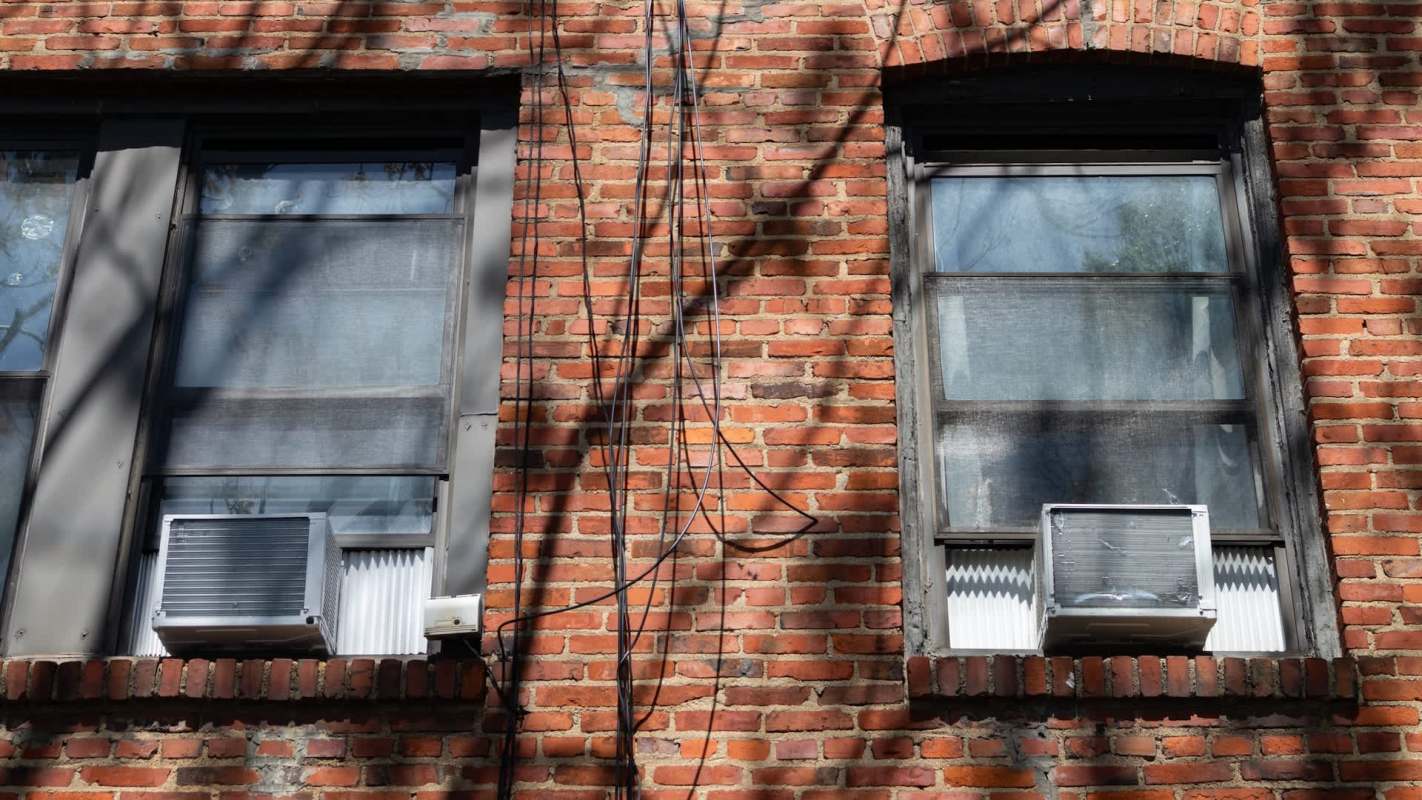 Two windows with air conditioners are set in a brick wall, with electrical wires running nearby.