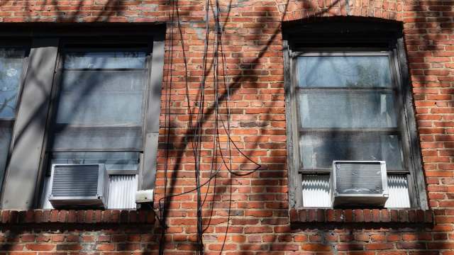 Two windows with air conditioners are set in a brick wall, with electrical wires running nearby.