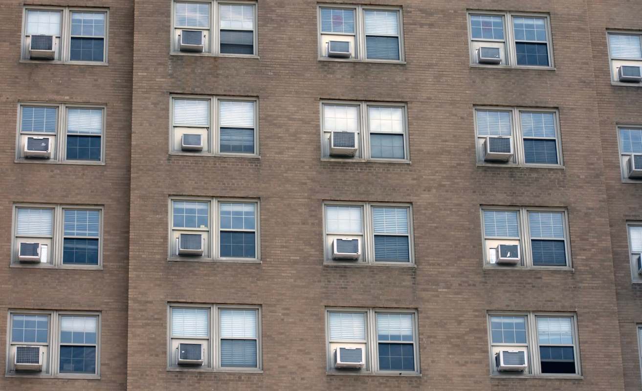 A brick building facade with several windows, each featuring air conditioning units.