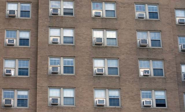 A brick building facade with several windows, each featuring air conditioning units.