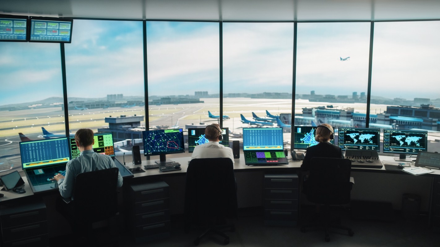 Air traffic control room with multiple monitors displaying flight data and a view of an airport runway.