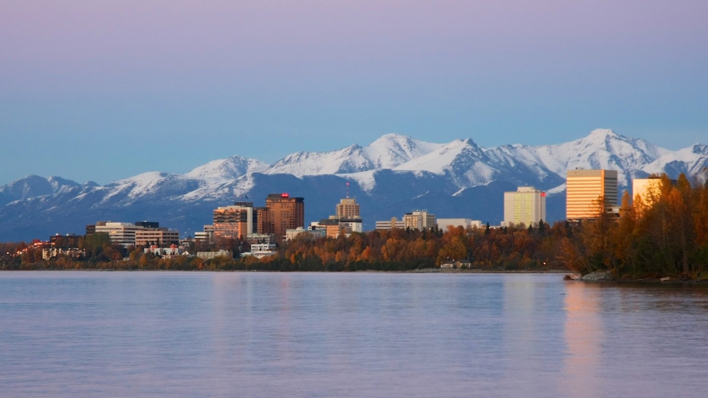 A city skyline with snow-capped mountains in the background and a calm body of water in the foreground.