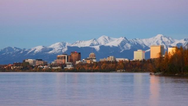 A city skyline with snow-capped mountains in the background and a calm body of water in the foreground.