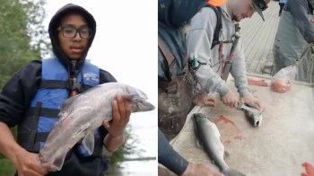 A person holds a fish, while another cuts fish on a table outdoors.