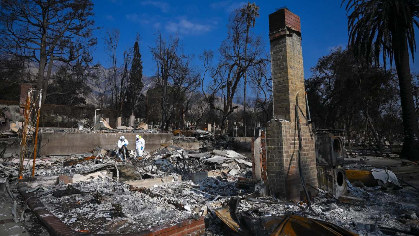 Two people in protective gear inspect the remains of a burned structure surrounded by charred trees and debris.